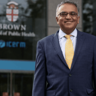 A man in a suit and tie standing in front of the brown school of public health.