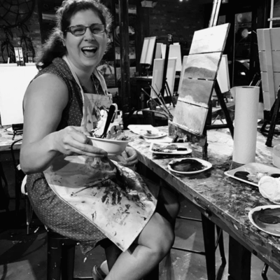 A woman in a black and white photo smiling at a painting table.
