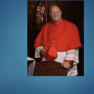 A priest in a red robe is posing in front of a blue background.