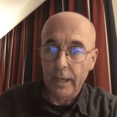 A bald man with glasses in front of a curtain.