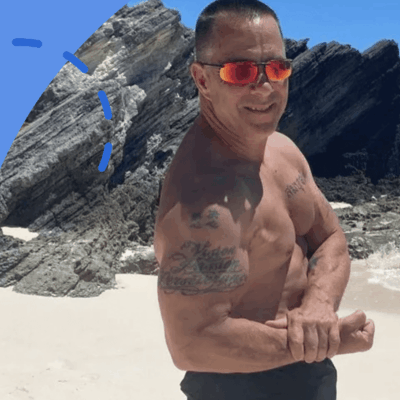 A shirtless man posing in front of a rocky beach.