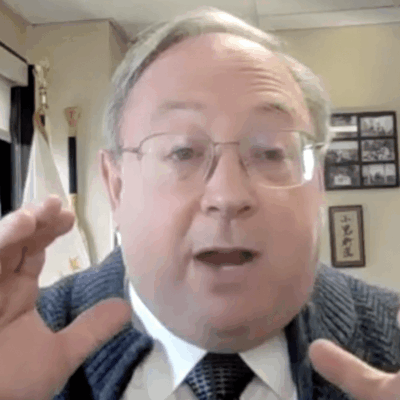 A man wearing glasses and a tie is speaking into a camera.