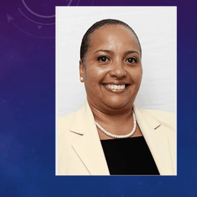 A woman in a blazer and pearls smiles in front of a purple background.