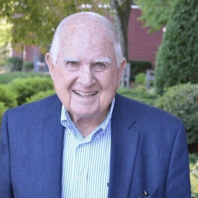 An older man in a blue suit smiles in front of bushes.