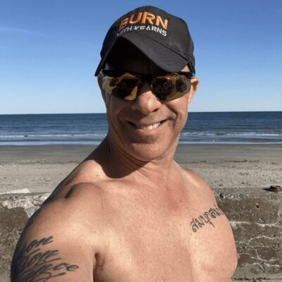 A man with tattoos and a hat in front of the ocean.