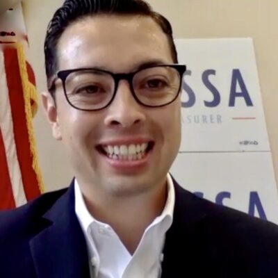 A man in glasses is smiling in front of an american flag.