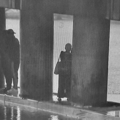 A black and white photo of a man standing under a bridge.