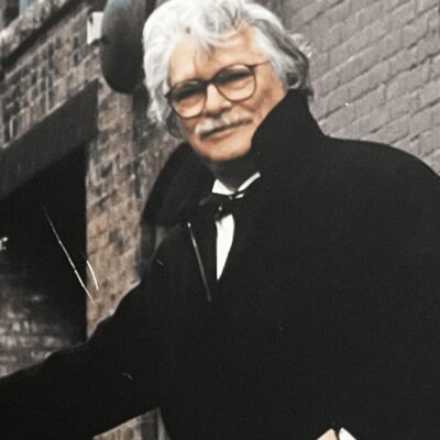 A man with glasses and a mustache standing in front of a brick building.