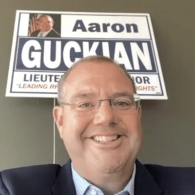 A man in a suit and tie is standing in front of a sign that says aaron guccian.