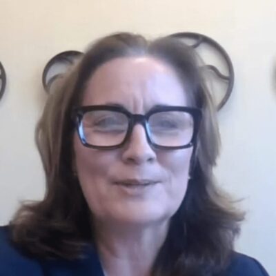 A woman wearing glasses and a suit in front of a wall.