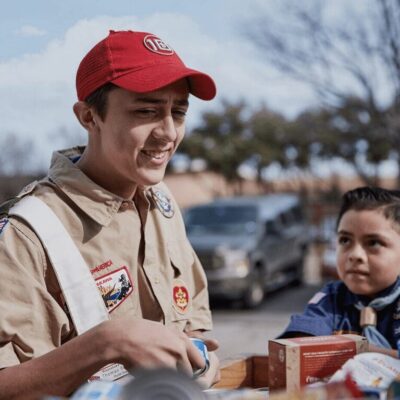 A boy in a scout uniform is standing next to a box of food.
