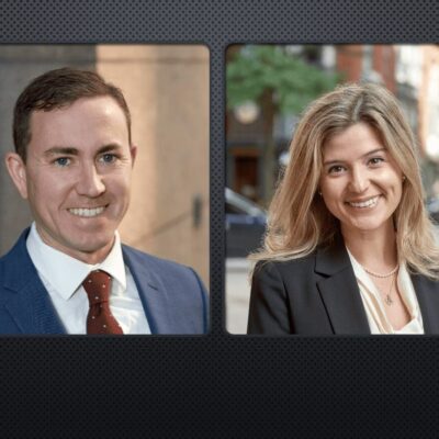Two people in business suits are smiling in front of a black background.