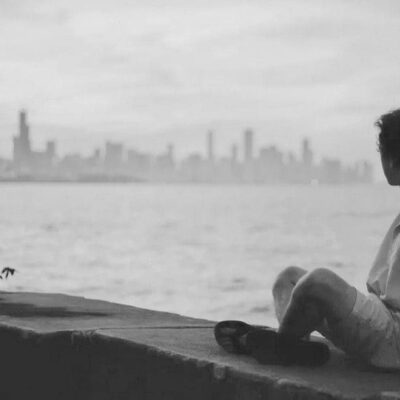 A man sitting on a wall looking at the water with a city skyline in the background, promoting inclusivity and equality.