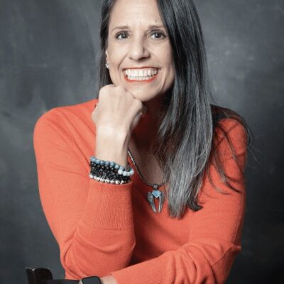 A smiling woman in an orange sweater sitting on a chair.
