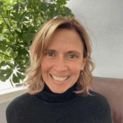 A smiling woman in a black turtleneck sitting in front of a potted plant at Social Enterprise Greenhouse.