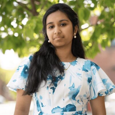 A young woman who identifies as BIPOC in a blue and white floral dress standing in front of a tree.