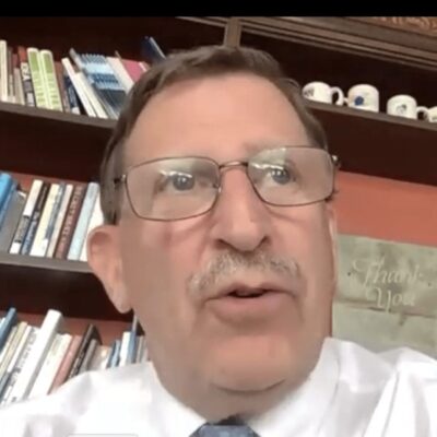A man wearing glasses and a tie proudly stands in front of a bookcase, symbolizing his affiliation with the Rhode Island Foundation.