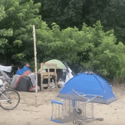 A group of tents and bicycles are parked in a dirt area occupied by homeless individuals.