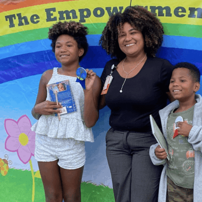 A woman and two children stand in front of a banner that says the Pawtucket Arts Festival's empowerment fund.