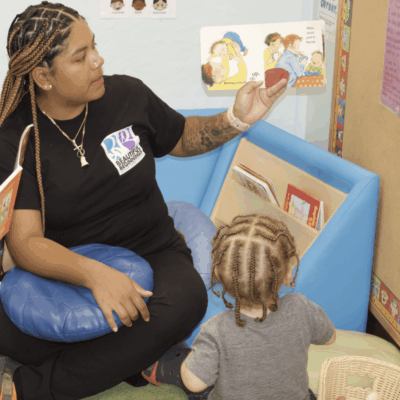 A woman providing food assistance to a child in a classroom while reading to them.