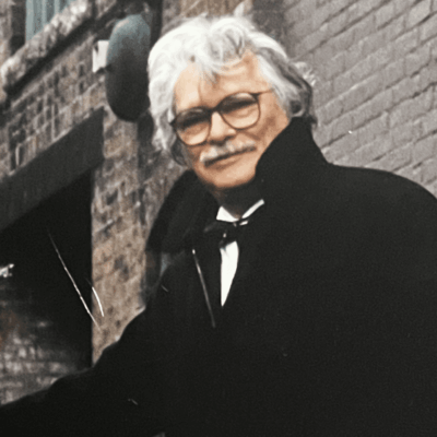 A man with glasses and a mustache is leaning against a brick wall.