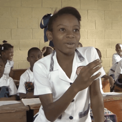 A girl from Haiti is standing in front of a classroom.