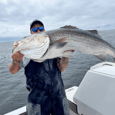 A man proudly displaying a massive striped bass while out on a boat in the picturesque outdoors of Rhode Island.