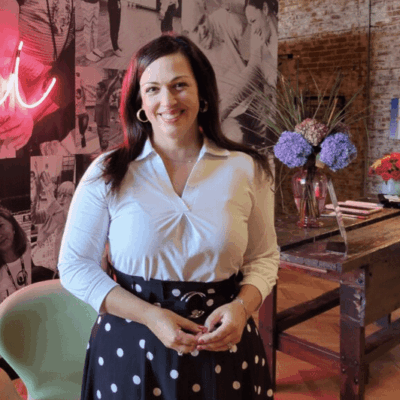 Alison Bologna, a woman in a polka dot skirt, standing in front of a neon sign.