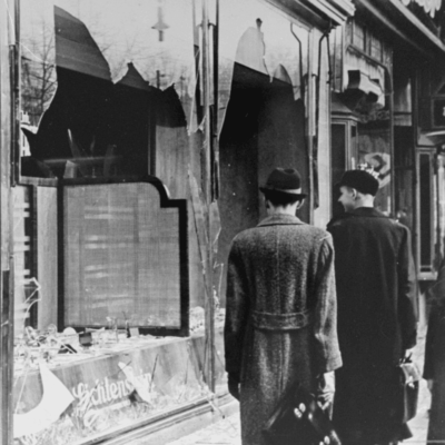 A group of people walking down a street during Kristallnacht with a broken window.
