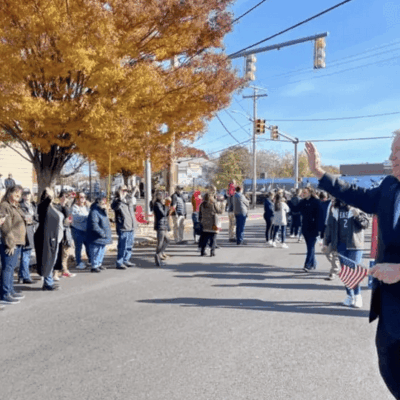 A man in a suit is waving to a crowd of people at the Cranston Veterans Day Parade.