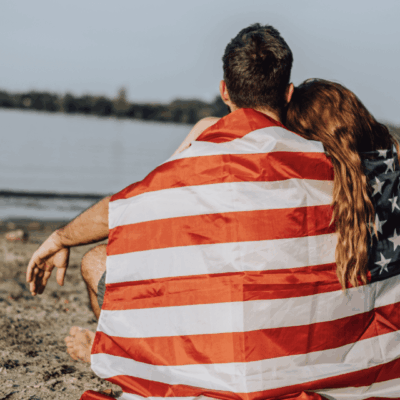 A patriotic couple embracing on the beach, enveloped in an American flag.