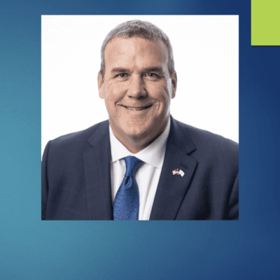 Greg Amore, in a suit and tie, is smiling in front of a blue background.