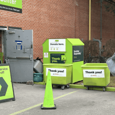 A woman is standing next to a recycling bin, donated used clothing for charity, in front of a building.