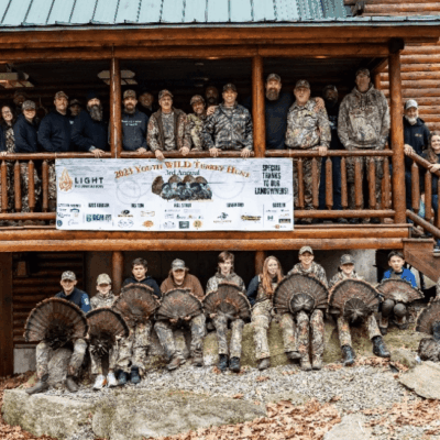 A group of people posing in front of a cabin before a turkey shoot for youth.