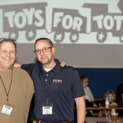 Two men standing next to each other in front of a toys for tots sign.