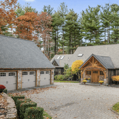 Rustic cabin with a porch next to a large garage surrounded by trees.