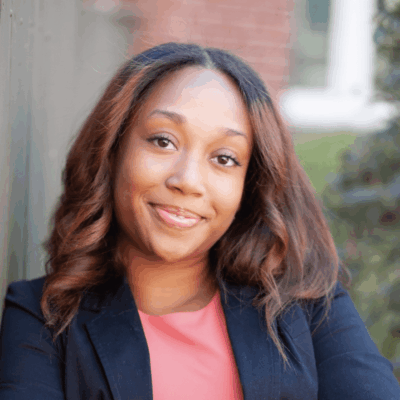 A smiling woman with brown hair wearing a black blazer and pink top.
