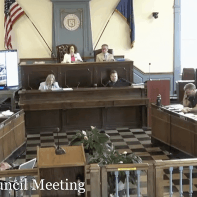 City council meeting in a formal chamber with members seated and American flags displayed.