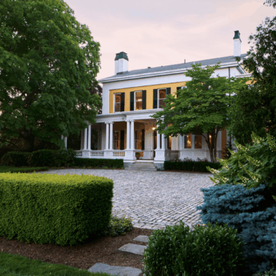 Elegant white mansion with columns surrounded by lush greenery and a cobblestone driveway.