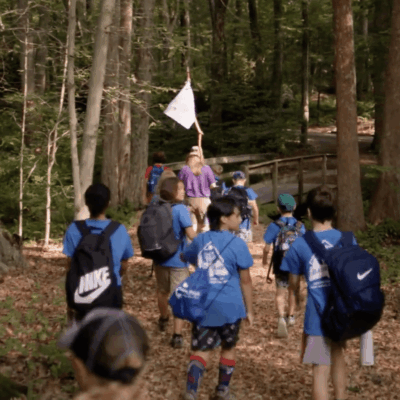 Children hiking on a forest trail, led by an adult with a white flag.