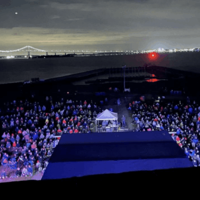 Large crowd gathered at an outdoor nighttime event with city lights in the background.