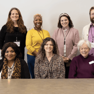 A diverse group of eight adults smiling for a group photo indoors.