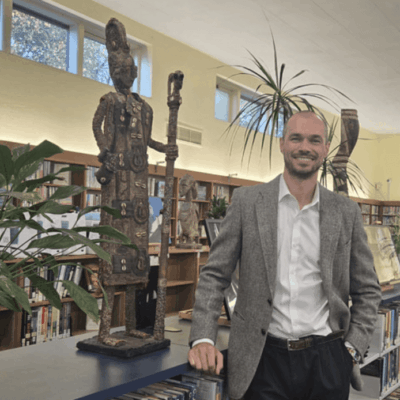 A man in a suit stands beside a tall wooden sculpture in a library.