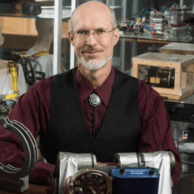 An older man in a vest and bolo tie in a workshop with mechanical devices.