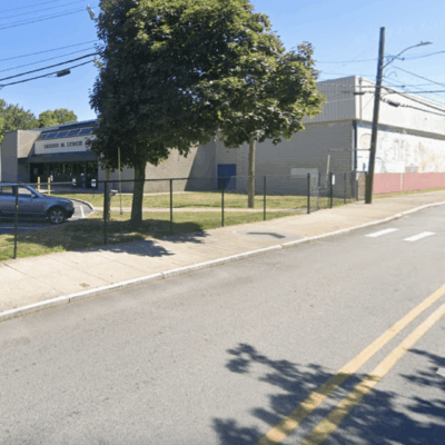 Street view of a fenced industrial building with parked cars and trees.