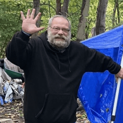 Man in black hoodie waving outdoors near a blue tarp and campsite.