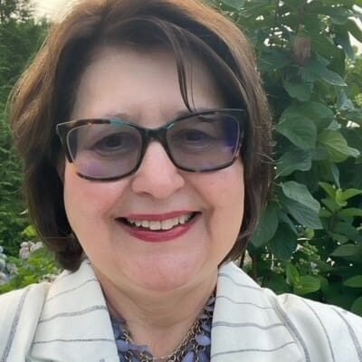 Woman smiling, wearing glasses and necklace.