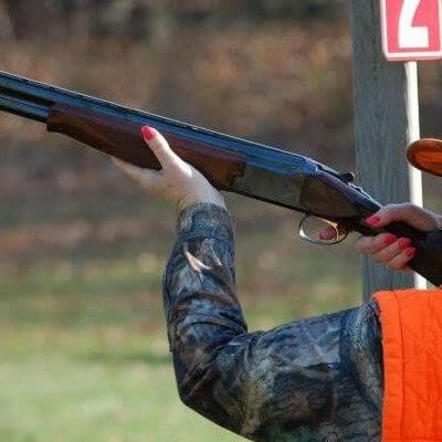 A woman wearing an orange hat and orange vest shooting a shotgun.