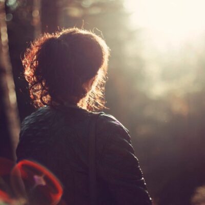 A woman is standing in the woods looking at the sun.