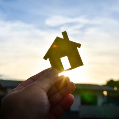 A person holding a house model in front of a sunset.